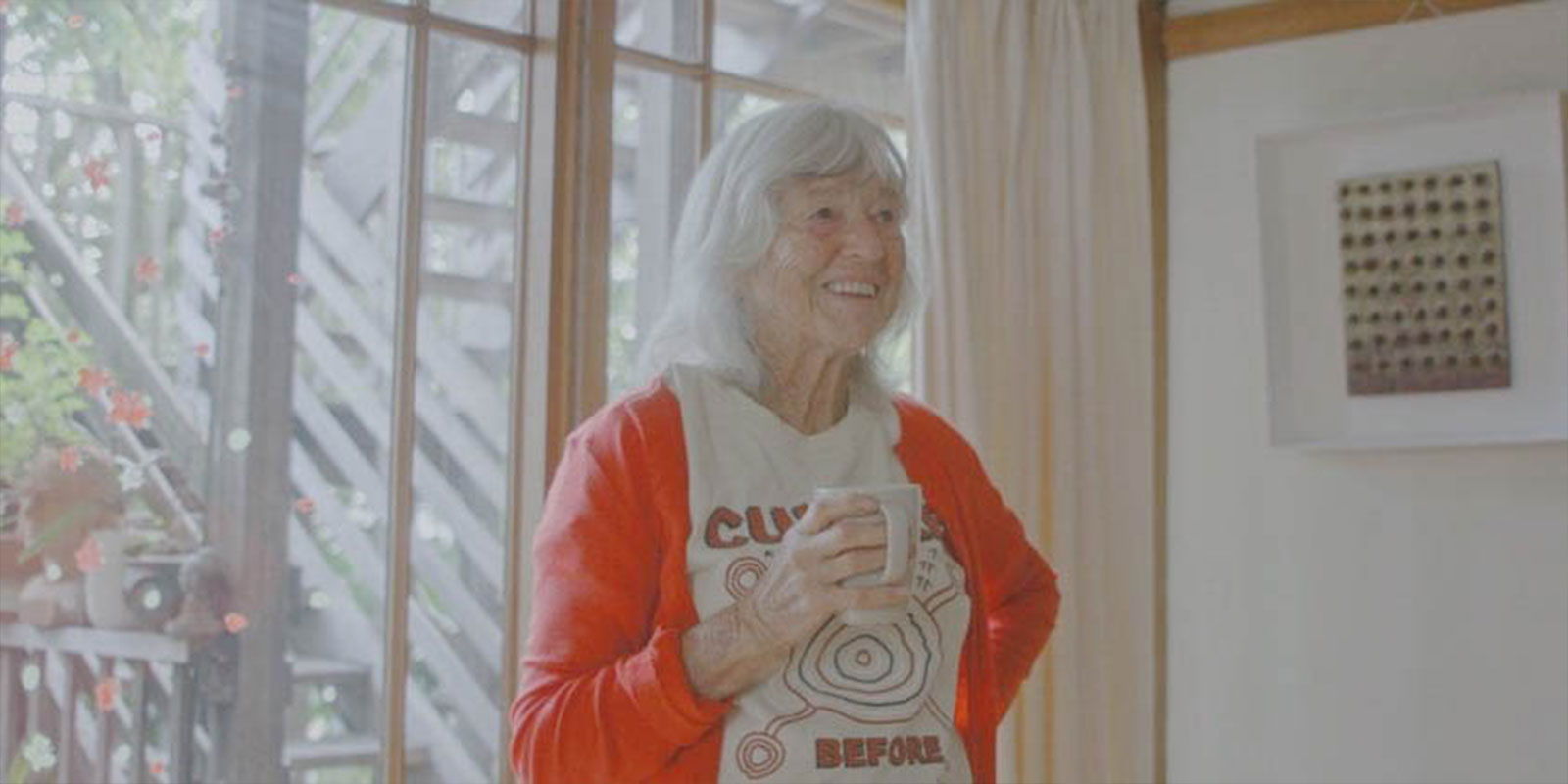 Image taken from Australian Short screening 'Save Our Soul', directed by Chloë Beckley. It features a silver-haired woman in a red cardigan smiling with a cup of tea